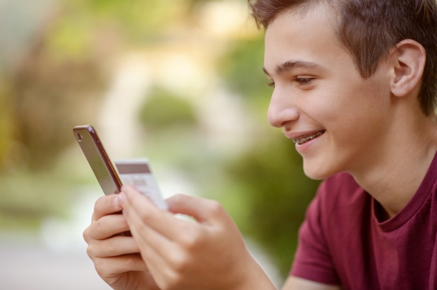 Teenage boy with a credit card and phone makes purchasing outdoors. Happy young man is using smart phone and bank card for online shopping. Handsome smiling white guy holds bank  card and cell phone.